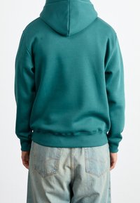 Hoodie in cotone turchese con cappuccio regolabile, polsini e orlo a costine, abbinato a pantaloni in denim azzurri chiari con tasche posteriori.