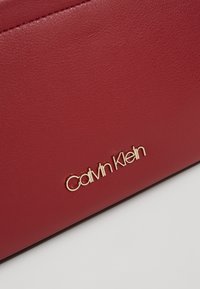 Czerwona, teksturowana skórzana powierzchnia z metalicznym, złotym logo Calvin Klein przymocowanym blisko dolnej krawędzi.