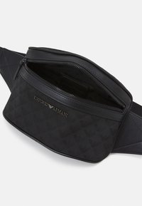 Černá ledvinka z texturované tkaniny, s logem, zipovým uzávěrem a nastavitelným popruhem. Interiér obsahuje jednu přihrádku.