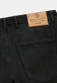 Jean en denim noir avec une poche arrière et une étiquette en cuir beige portant l'inscription "Serge Blanco, Celui qui va à tout le monde, Depuis 1958"