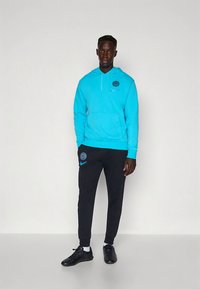 Sweatshirt azul claro com capuz, bolso frontal e detalhes a preto, combinado com calças de treino pretas com um logótipo. Tênis desportivos pretos completam o conjunto.