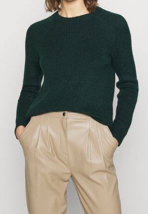 Femme portant un pull côtelé vert foncé rentré dans un pantalon en cuir plissé beige, les mains dans les poches, vue du cou à mi-cuisse.