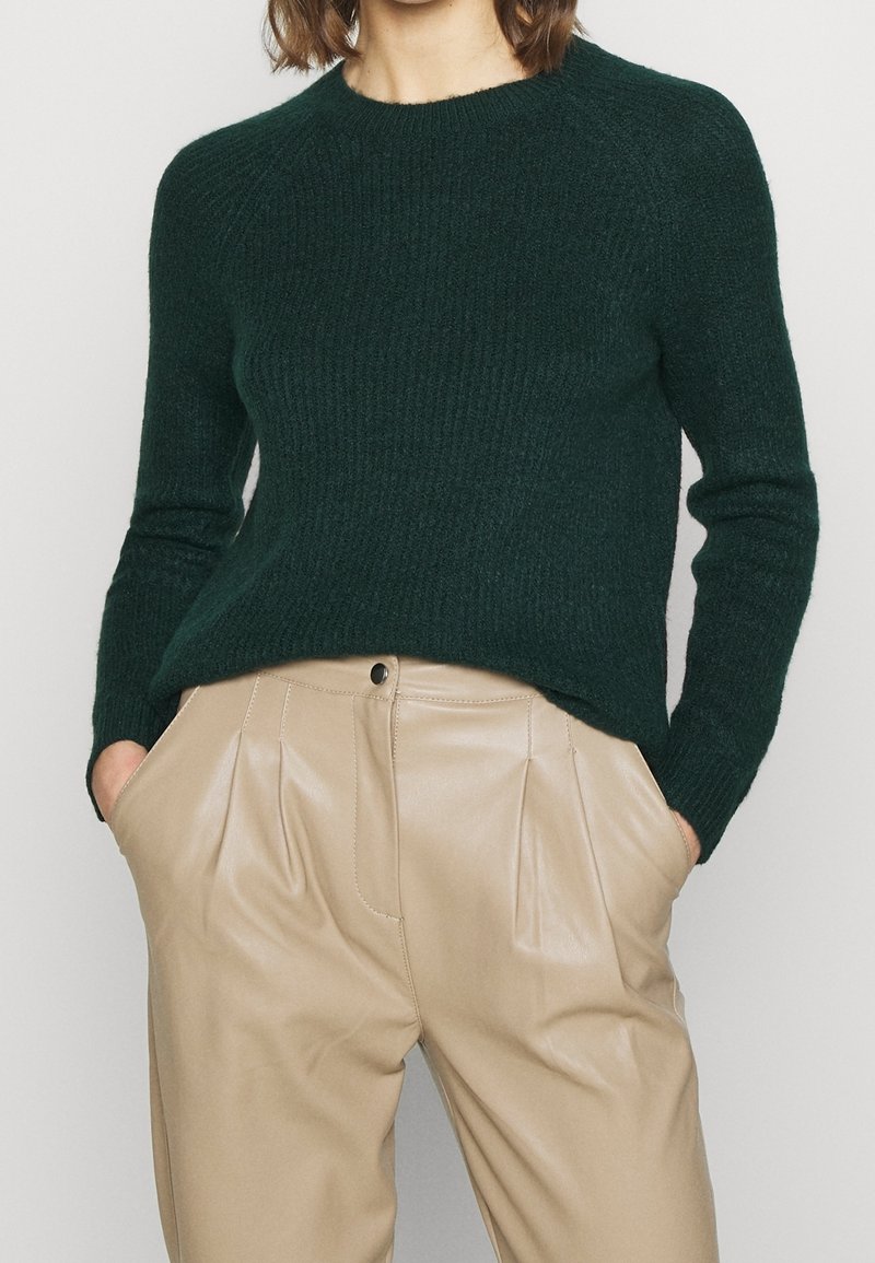 Femme portant un pull côtelé vert foncé rentré dans un pantalon en cuir plissé beige, les mains dans les poches, vue du cou à mi-cuisse.