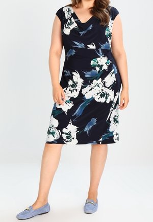Mujer con un vestido negro hasta la rodilla con grandes estampados florales blancos y verdes y zapatos tipo slip-on azul claro.