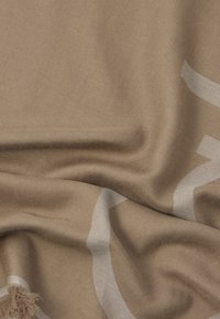 Beige vævet stof med subtile hvide buede linjer og bløde folder, som viser tekstur og frynset kant.