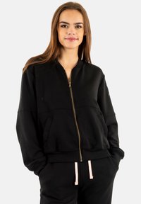 Sudadera negra con cremallera, cuello redondo, mangas largas y dos bolsillos frontales; cuenta con cordones rosa contrastantes en pantalones a juego.