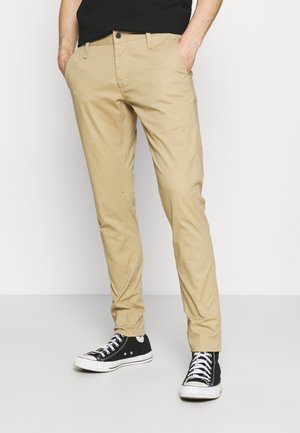 Man staat met handen in de zakken, draagt beige casual broek, zwarte hoge sneakers met witte veters en een zwart shirt.