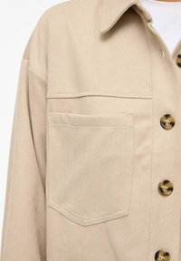 Bēša kokvilnas krekla jaka ar klasisku apkakli, priekšējo kabatu un apaļiem misiņa pogām, kas demonstrē gludu, teksturētu virsmu.