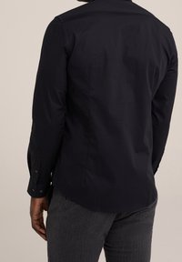 Muž v černém přiléhavém triku s dlouhým rukávem a šedých kalhotách, zobrazený zezadu na jednobarevném pozadí.