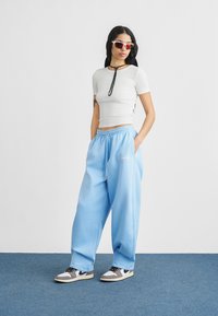 Γυναίκα που φοράει λευκό εφαρμοστό κοντό μπλουζάκι, ανοιχτό μπλε φαρδιά φόρμα, αθλητικά παπούτσια και γυαλιά ηλίου με κόκκινα κρύσταλλα, στέκεται σε μπλε μοκέτα μπροστά σε λευκό τοίχο.