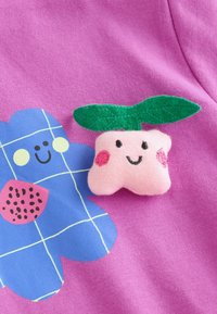 Lyserødt stof med en lille blød bamse formet som en smilende lyserød frugt med grønne blade, ved siden af et blåt smilende blomstermotiv med ternet mønster.