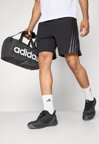 Schwarze Sportshorts mit seitlichen Streifen, kombiniert mit schwarzen Sportschuhen und weißen Socken mit einem schwarzen Logo, sowie einer grauen Sporttasche.
