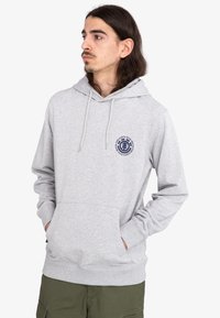 Sudadera gris claro con un bolsillo frontal, capucha con cordón y un logo circular en azul marino en el pecho. Presenta puños y dobladillo acanalados.