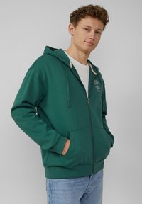 Grüne Kapuzensweatjacke mit einem Kordelzug, frontalen Taschen und gerippten Bündchen. Mit einem Logo auf der Brust. Über einem weißen Shirt getragen.