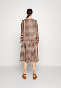 Hnědé vzorované šaty s dlouhým rukávem obsahující houndstooth a pruhované akcenty. Rozšířená sukně a lehký materiál pro pohodlí. Viditelné ploché sandály.