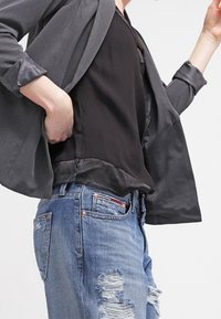 Osoba w szarym żakiecie noszącym czarną satynową bluzkę i przetarte niebieskie dżinsy, dostosowująca żakiet, trzymając jedną rękę w kieszeni.