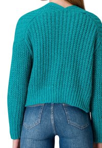 Türkiser gestrickter Pullover mit texturiertem Finish und geripptem Saum. Der verkürzte Schnitt betont die Rückendetails. Kombiniert mit blauer Jeans.