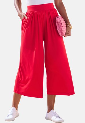 Woman wearing bright red wide-leg cropped pants, white sneakers, and holding a pink textured clutch with one hand in pocket.