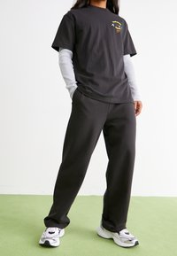 Osoba oblečená v černém tričku, pod ním má dlouhé pruhované bílé rukávy, černé kalhoty a bílé tenisky, stojí na světle zelené podlaze.