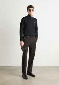 Crni džemper sa patentnim zatvaračem, smeđe krojene hlače, crni remen, drži crnu torbicu, nosi velike sunčane naočale, stoji na betonskoj podlozi.