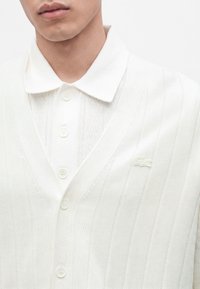 Gros plan d'une personne portant un polo blanc boutonné sous un cardigan blanc côtelé avec un logo brodé subtil sur la poitrine.