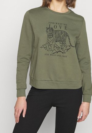 Femme portant un sweat-shirt vert olive avec un graphique de tigre noir et le texte "Trouve ce que tu aimes, reste sauvage et libre", accompagné d'un pantalon noir.