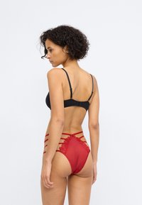 Schwarzer BH mit verstellbaren Trägern und roter hochgeschnittener Panty, die mit Spitze und Mesh-Verzierungen ausgestattet ist, mit horizontalen Trägern auf der Rückseite.
