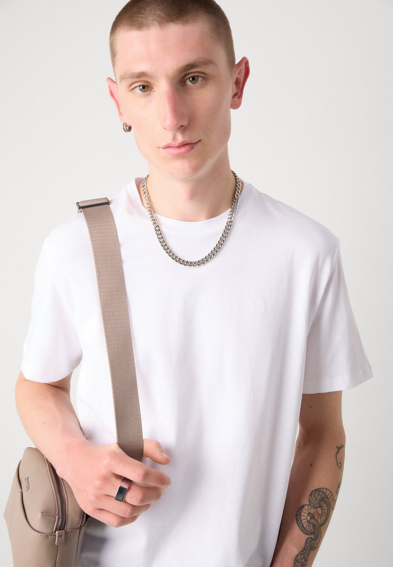 Biała koszulka z krótkim rękawem z subtelnym logo, noszona z srebrnym łańcuszkiem na szyi. Widoczna jest również beżowa torba na ramię i ciemny pierścionek.