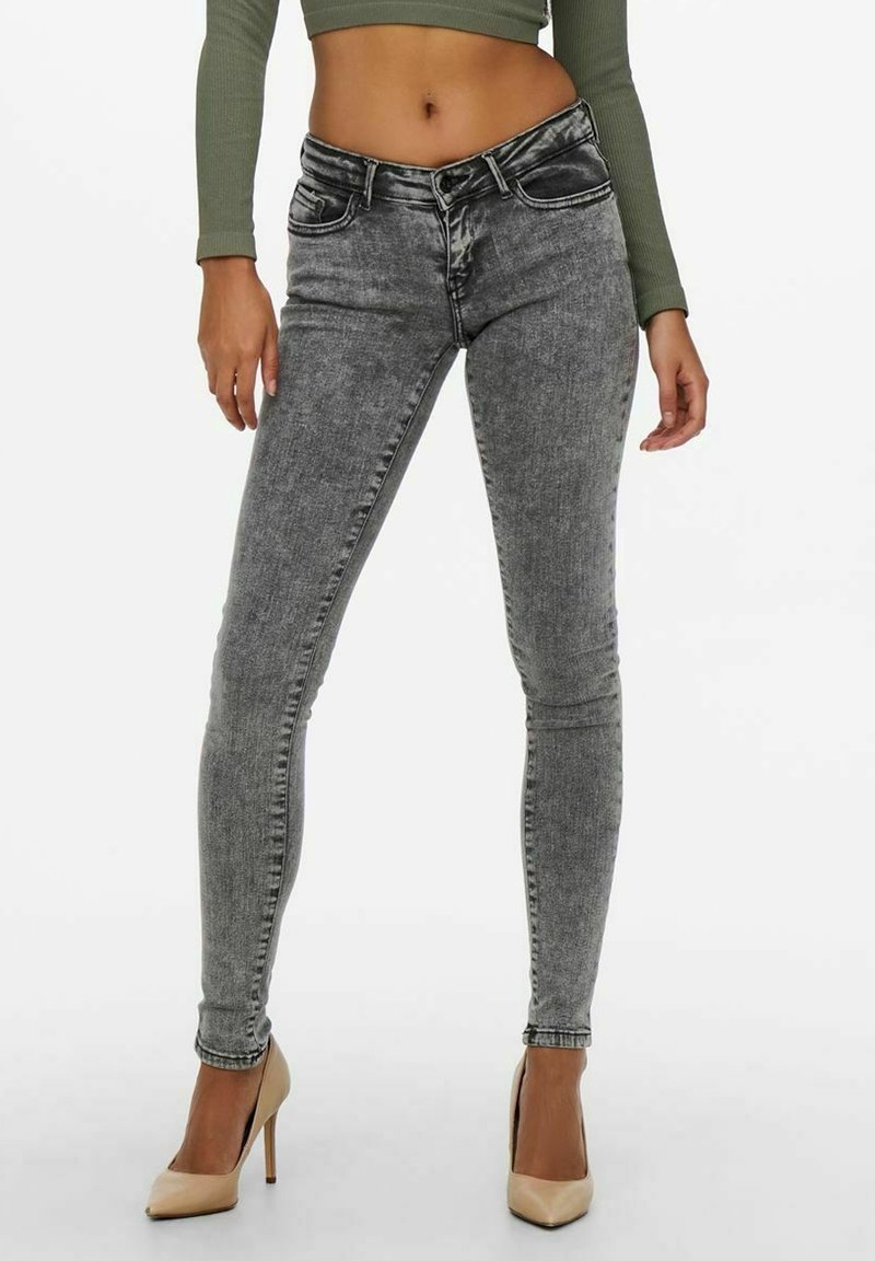 Jeans skinny gris avec une finition délavée, un design à cinq poches et des détails de couture subtils, associés à des talons hauts beiges et un haut vert ajusté.