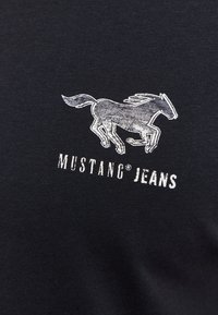 Czarna t-shirt z materiału z logo konia w kolorze srebrnym wytłoczonym nad napisem "MUSTANG® JEANS" w białym kolorze. Gładka tekstura.