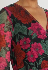 Blumenkleid mit tiefem V-Ausschnitt, das große rote und pinke Blumen auf einem schwarzen, durchsichtigen Stoff zeigt. Grüne Blätter betonen das Design.