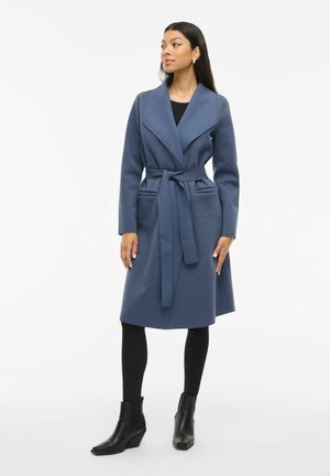 Langer, blauer Wickelmantel mit einem Gürtel, breitem Kragen und Fronttaschen. Getragen über schwarzen Leggings und Stiefeletten. Glatte Textur.