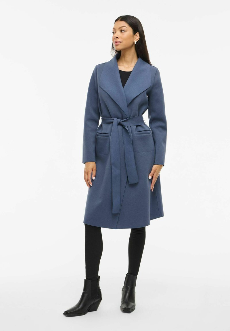 Cappotto avvolgente blu lungo con cintura, caratterizzato da un ampio colletto e tasche frontali. Indossato sopra leggings neri e stivaletti alla caviglia. Texture liscia.