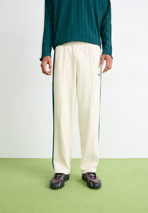 Cream-colored athletic pants with dark green side stripes and an elastic waistband, paired with black and green sneakers. Textured fabric design.