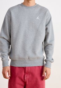 Szary sweter z okrągłym dekoltem, z prążkowanymi mankietami i dołem, z małym białym logo na klatce piersiowej. Noszony z czerwonymi spodniami.