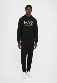 Fekete kapucnis pulóver és melegítőnadrág szett, arany EA7 Emporio Armani logóval. Puha anyagból készült, bordás mandzsettákkal és elülső zsebbel, fehér sportcipővel párosítva.