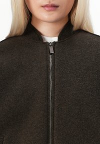 Brun, ullblandet jakke med høy krage og glidelås. Teksturen er myk, og designet er minimalistisk, med fokus på varme og komfort.