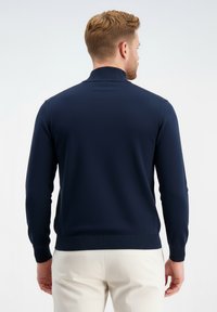 Vyras su trumpais šviesiai rudos spalvos plaukais, dėvintis tamsiai mėlyną megztinį ilgomis rankovėmis ir smėlio spalvos kelnes, stovintis nugara balto fono atžvilgiu.