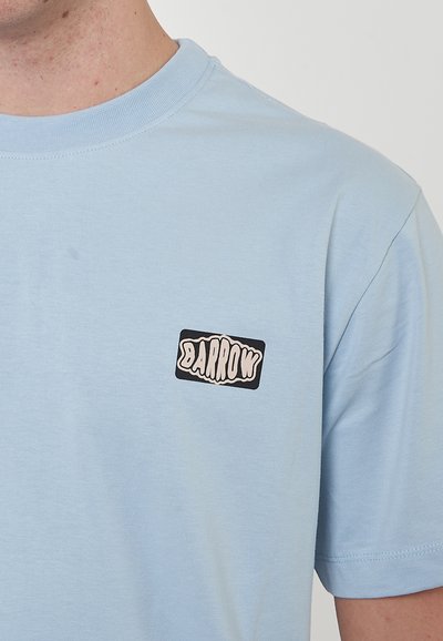 Camiseta de algodón azul claro con cuello redondo. Presenta un pequeño parche rectangular negro con texto blanco que dice "BARROW" en la parte izquierda del pecho.