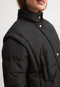 Must mustkunst, millel on kõrge krae, trukkide kinnitused ja tikitud tekstuur. Esiküljel on küljetasku.