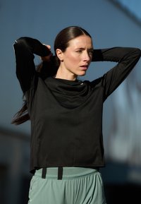 Crna majica dugih rukava s opuštenim krojem i vratom u obliku kapuljače, uparena s svijetlozelenim sportskim kratkim hlačama koje imaju elastični pojas i mrežastu podstavu.