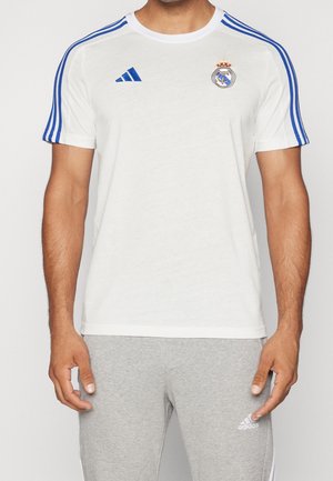 Mężczyzna w białej koszulce Realu Madryt z niebieskimi paskami Adidas na rękawach i szarych spodniach dresowych Adidas.