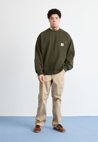Sudadera verde oscuro con bolsillo frontal y logo, combinada con pantalones beige y zapatillas marrones. Tela suave, corte casual, diseño minimalista.