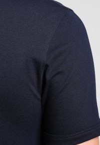 Detailní záběr na horní část paže osoby oblečené v námořnicky modrém tričku s krátkým rukávem, na jednobarevném šedém pozadí.