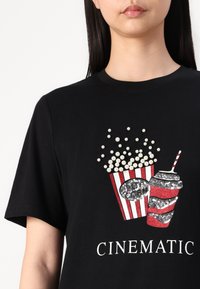 Zwarte katoenen t-shirt met een met sequins versierde afbeelding van popcorn in een gestreepte doos en een beker met een rietje, gelabeld "CINEMATIC."