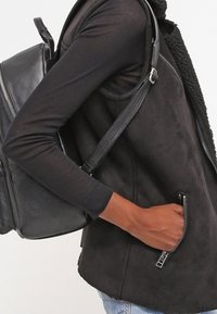 Persona che indossa una maglietta nera a maniche lunghe e un gilet, con una mano nella tasca con zip, mentre porta uno zaino di pelle nera su una spalla.