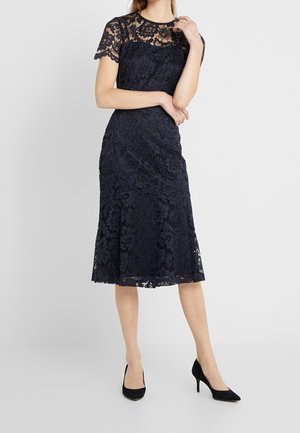 Vrouw die een knielange marineblauwe kanten jurk met korte mouwen en zwarte hoge hakken draagt, staand tegen een effen achtergrond.