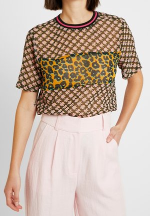 Frau trägt ein kurzärmeliges, gemustertes Mesh-Oberteil mit pink-braunem Kreuzmuster und einem breiten Leopardenmuster-Streifen, kombiniert mit hoch taillierter, hellrosa Hose.