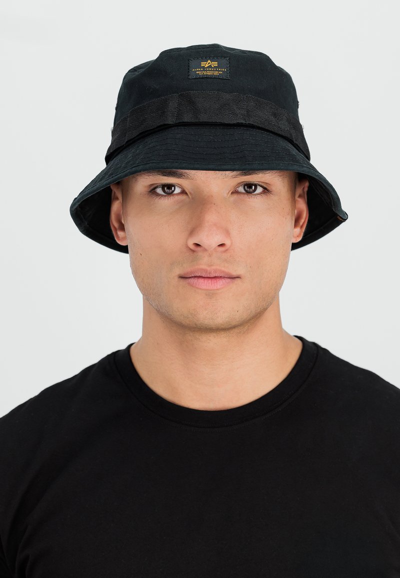 Jeune homme portant un chapeau seau noir avec un patch logo et un t-shirt noir à col rond, regardant droit devant sur un fond uni.