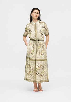 Žena stojaca, oblečená v krémovom midi šate so zelenými a hnedými vzormi vtákov a kvetov, krátkymi rukávmi, golierom a opaskom viazaným v páse.